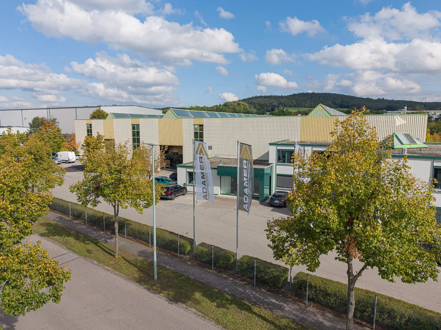 Hinter einem eingezäunten Gelände mit Bäumen und einem Gehweg steht ein modernes Industriegebäude mit grünen und beigen Platten. Drei große Fahnen mit der Aufschrift ADAME sind in der Nähe des Eingangs unter einem teilweise bewölkten Himmel angebracht.