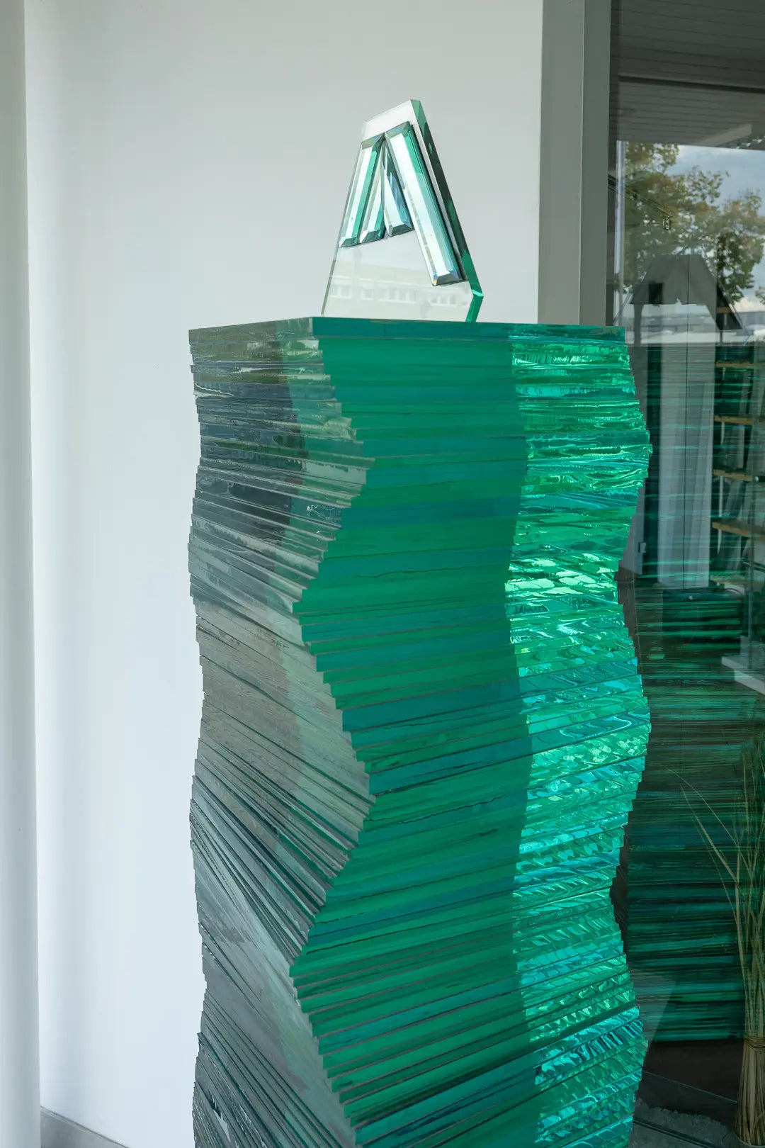 Eine hohe, abstrakte Skulptur aus übereinander gestapelten, gezackten grünen Glasplatten steht vor einem Fenster, reflektiert das Licht und erzeugt eine mehrschichtige Struktur. Ein kleineres dreieckiges Glasstück sitzt obenauf.