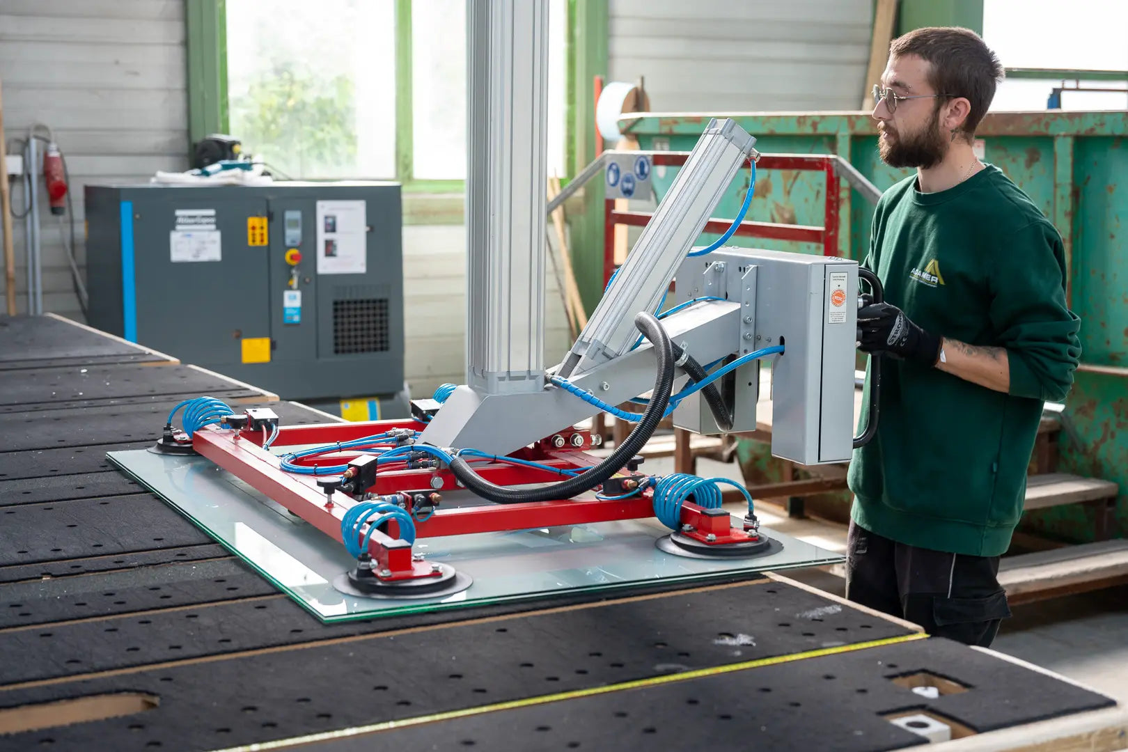 Ein Mann bedient eine Maschine mit Saugnäpfen zum Anheben und Bewegen einer Glasscheibe in einer Industriehalle. Er trägt Schutzhandschuhe und eine Schutzbrille. Im Hintergrund sind Industrieanlagen und ein großes grünes Gebäude zu sehen.