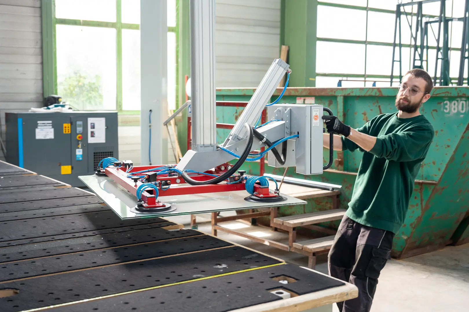 Ein Mann in einem grünen Hemd bedient eine Maschine mit Saugnäpfen, um eine große Glasscheibe in einer hell erleuchteten Industriehalle anzuheben.