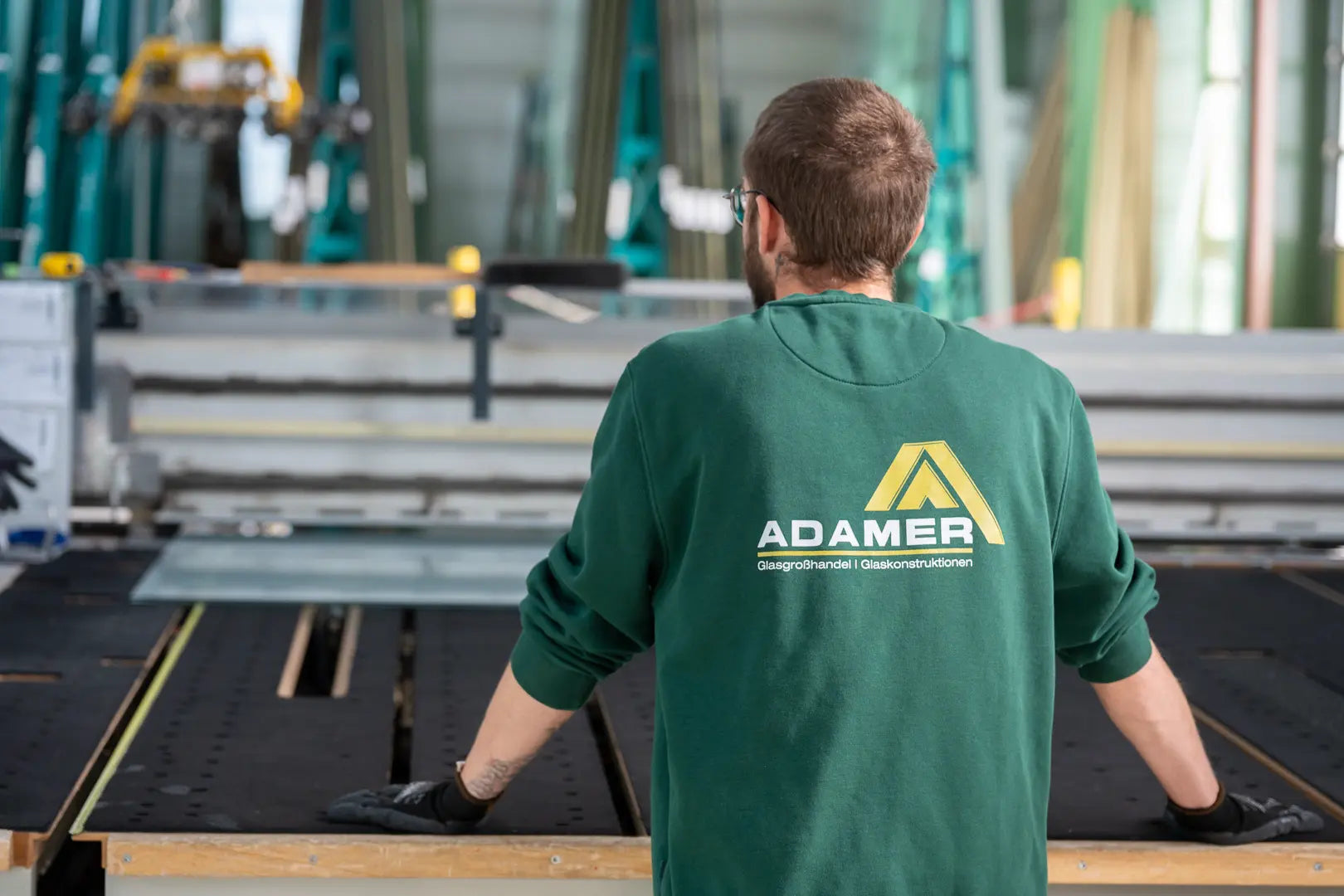 Ein Mann in einem grünen Hemd mit der Aufschrift ADAMER auf dem Rücken steht an einem Arbeitsplatz in einer Fabrik, vor Maschinen und Glasscheiben, die Hände auf den Tisch gestützt.