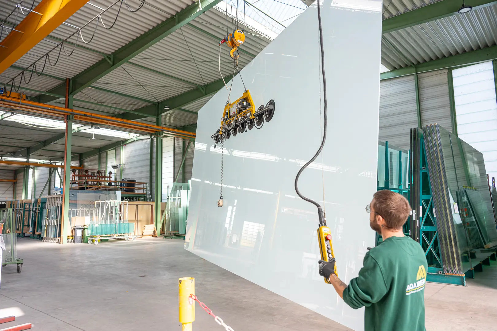 Ein Arbeiter bedient eine Maschine zum Anheben einer großen Glasscheibe mit einer Saugvorrichtung in einem geräumigen Industrielager, das mit Glasscheiben und Metallkonstruktionen gefüllt ist.
