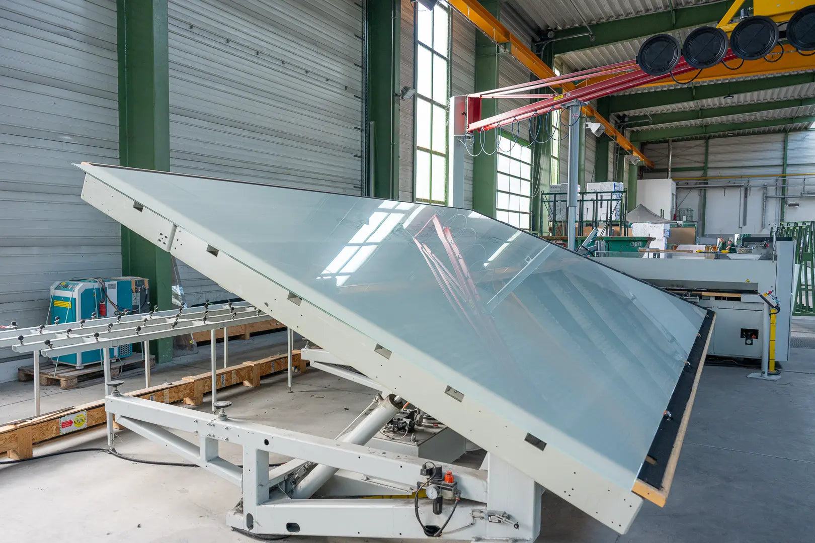 Große Glasscheibe auf einer gekippten, weißen Industriemaschine in einer geräumigen Fabrik mit hohen Decken, großen Fenstern und verschiedenen Geräten im Hintergrund.