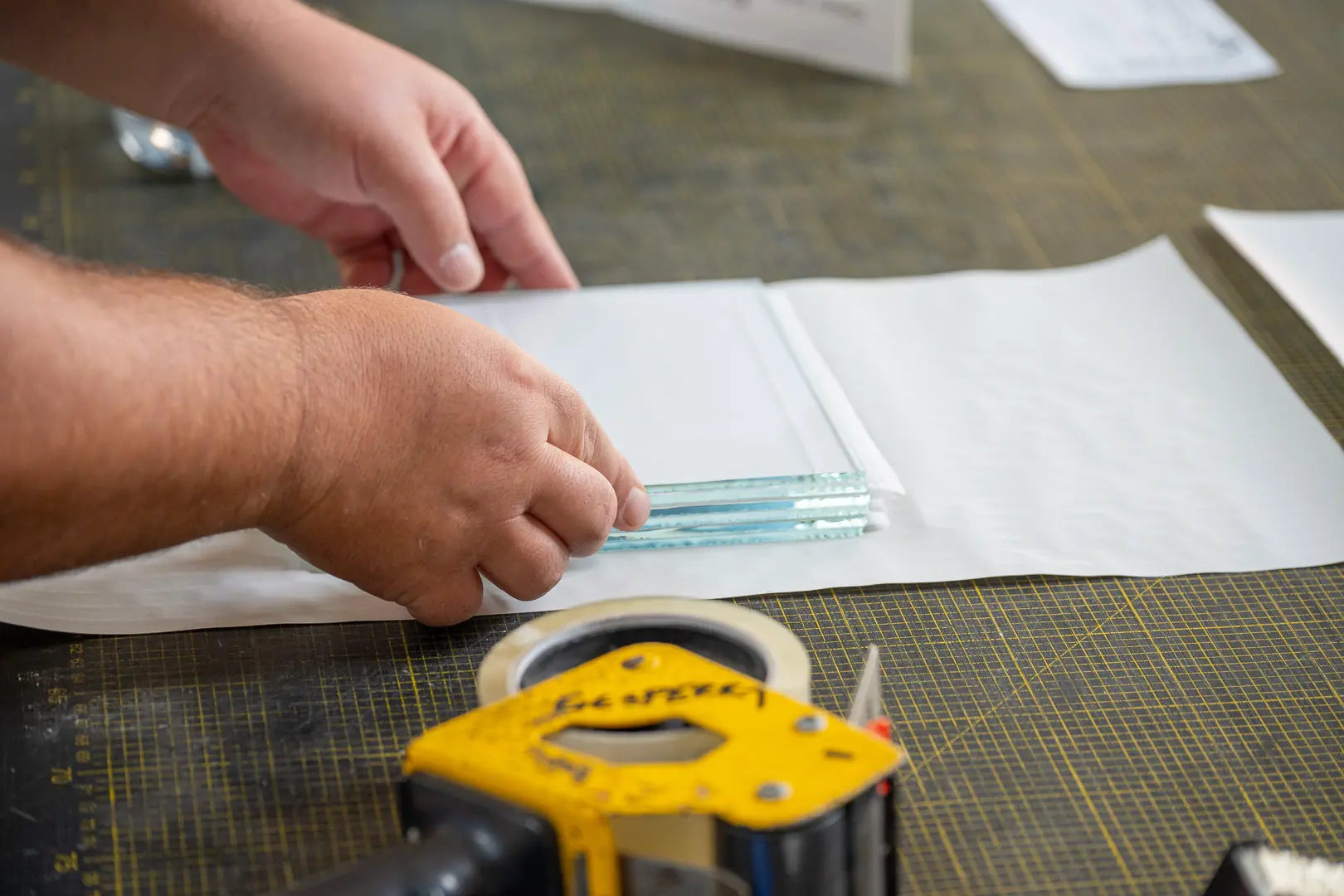 Die Hände einer Person wickeln auf einem Arbeitstisch drei kleine Glasscheiben sorgfältig in weißes Schutzmaterial ein, wobei durchsichtiges Klebeband und ein gelber Klebebandabroller in der Nähe sind.