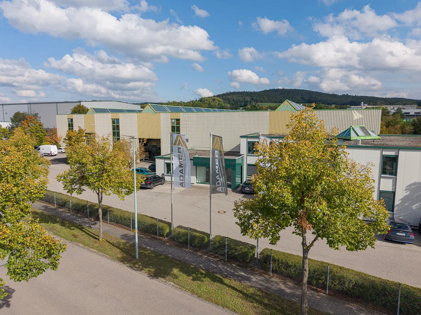 Luftaufnahme eines modernen Industriegebäudes mit grünen und beigen Akzenten, gesäumt von Bäumen und geparkten Autos im Freien. Drei vertikale Fahnen sind in der Nähe des Eingangs angebracht, und über dem Gebäude ist ein blauer Himmel mit Wolken zu sehen.