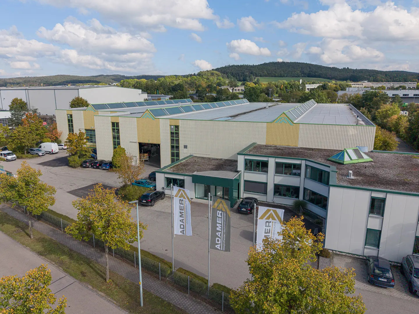 Ein modernes Industriegebäude mit großen Fenstern und grüner Verkleidung, umgeben von Bäumen und geparkten Autos. Vor dem Gebäude sind mehrere ADAMER-Flaggen angebracht, im Hintergrund sind Hügel und blauer Himmel zu sehen.
