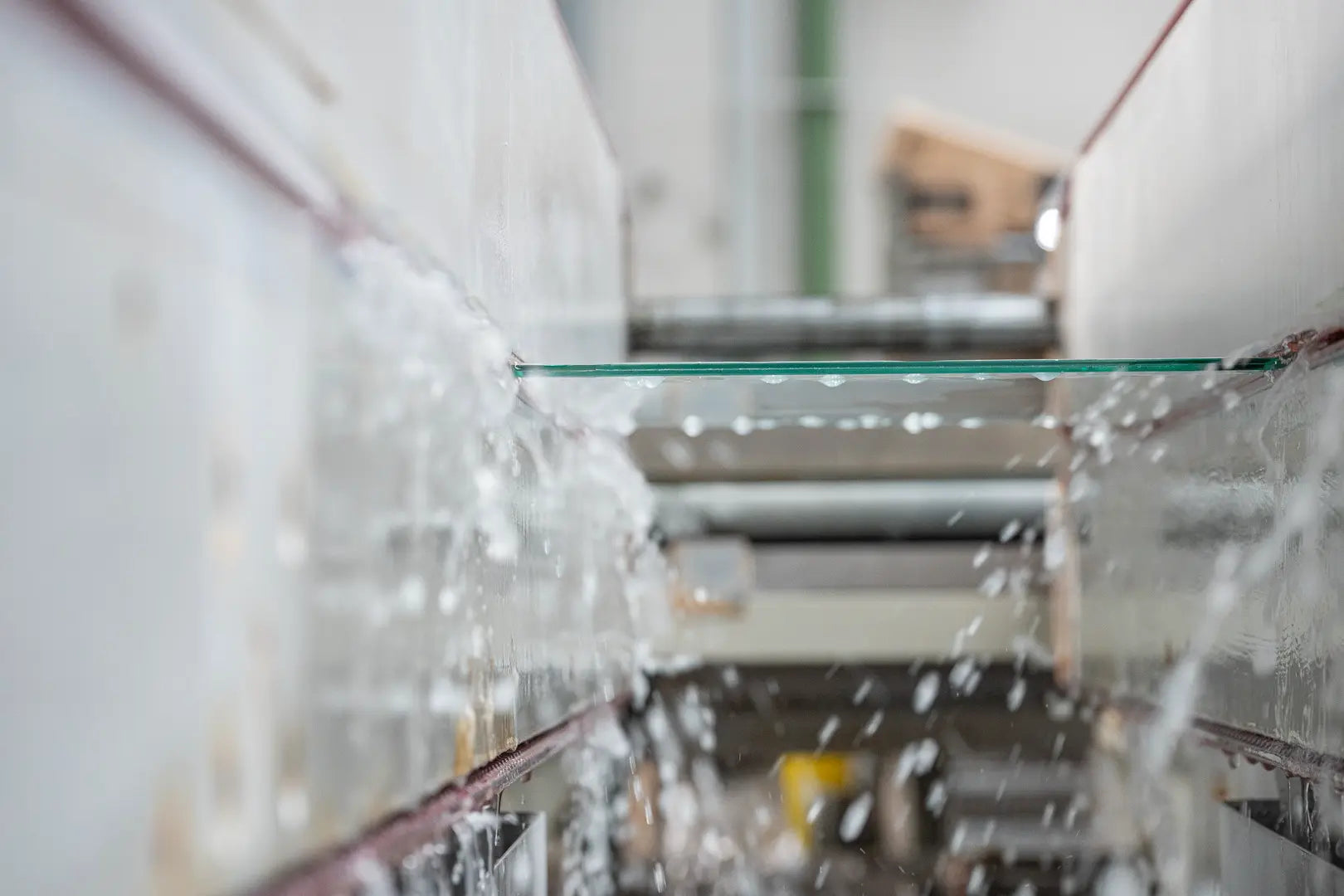 Eine Glasscheibe bewegt sich auf einem Förderband, während sie in einer industriellen Umgebung von beiden Seiten mit Wasserstrahlen besprüht wird. Der Hintergrund ist unscharf und konzentriert sich auf das Glas und das Wasser.