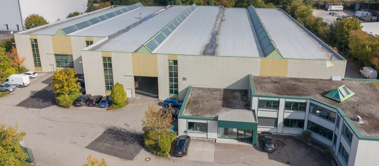 Luftaufnahme eines großen Industrielagers mit grauem Dach, Oberlichtern, Ladetoren und einem angebauten Bürogebäude, vor dem Autos geparkt sind und Bäume das Grundstück umgeben.