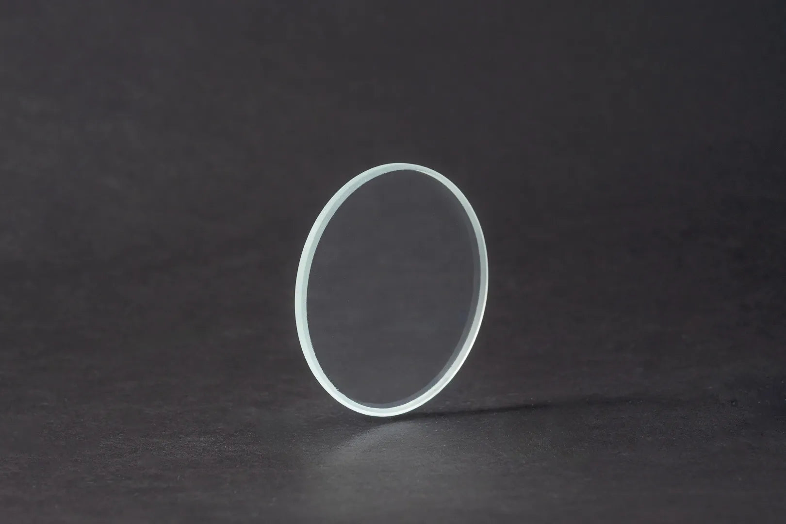 Eine einzelne, dünne, runde Platte aus Entspiegeltes Glas steht aufrecht auf einer dunklen, glatten Oberfläche mit einer subtilen Reflexion darunter.