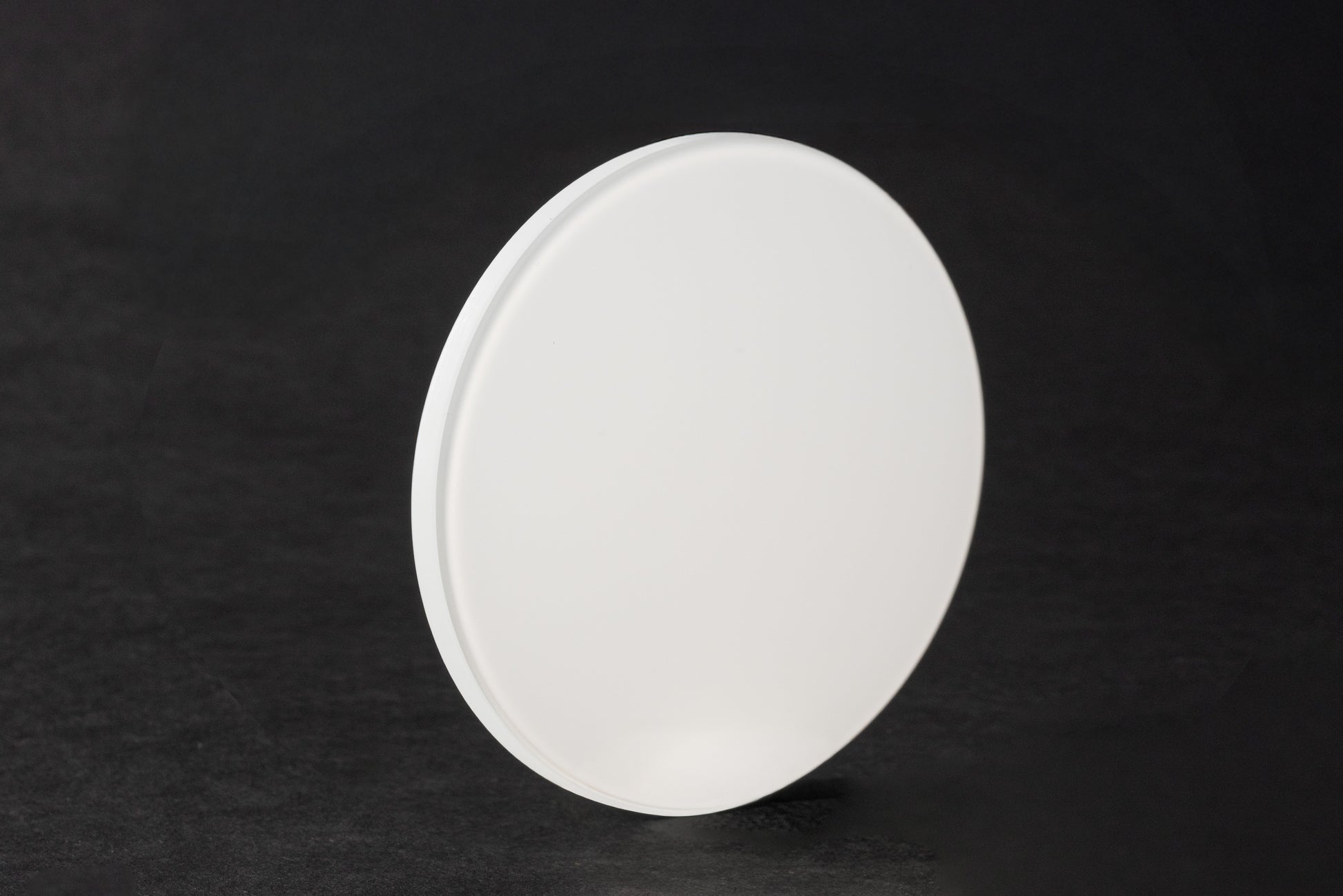 Ein rundes, flaches Milchüberfangglas steht aufrecht auf einer dunklen, strukturierten Oberfläche mit sanfter Beleuchtung vor einem schwarzen Hintergrund.
