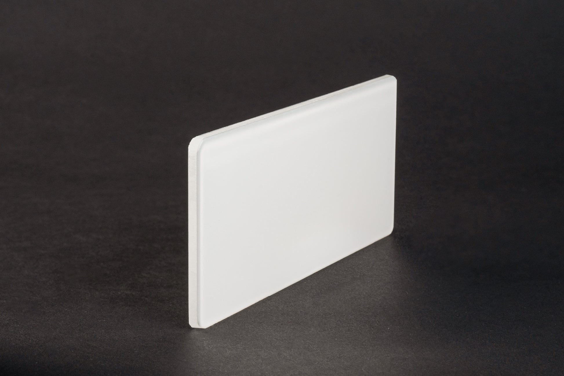 Das Milchüberfangglas ist eine rechteckige, mattierte weiße Acrylplatte mit glatten Kanten, die auf einer dunklen Fläche aufrecht steht. Sein halbtransparentes Design sorgt für eine diffuse Beleuchtung und ähnelt dem klassischen Milchüberfangglas.