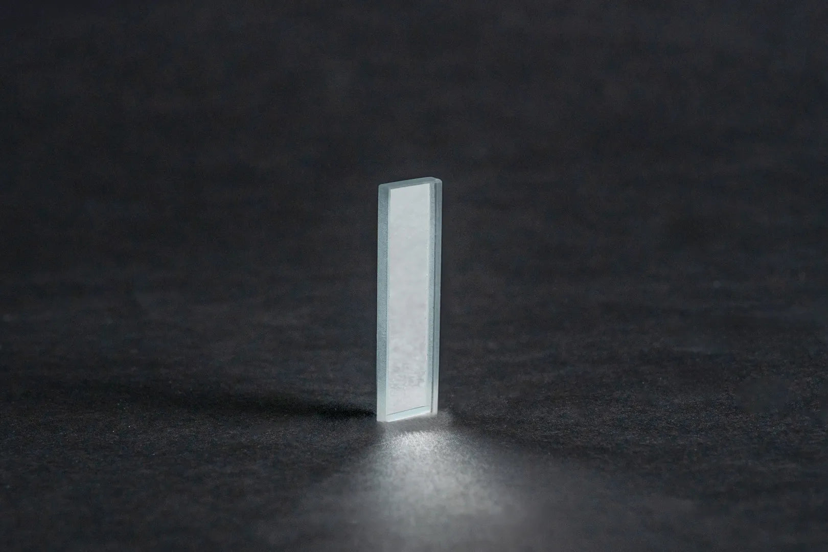 Eine rechteckige, transparente Oberflächenspiegel-Scheibe (Weißglas-Basis) steht aufrecht auf einer dunklen, strukturierten Oberfläche und wirft einen schwachen Schatten und eine subtile Reflexion unter sich.