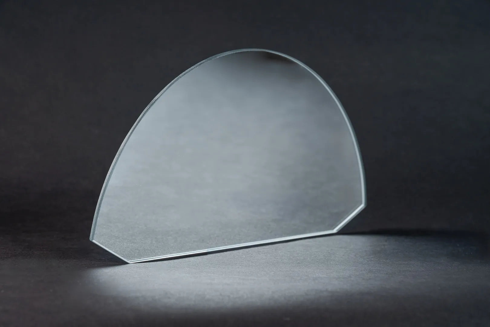 Ein dünner, transparenter, halbkreisförmiger Oberflächenspiegel (Weißglas-Basis) steht aufrecht auf einer dunklen Fläche vor einem dunklen Hintergrund und wirft eine subtile Reflexion und Schatten.