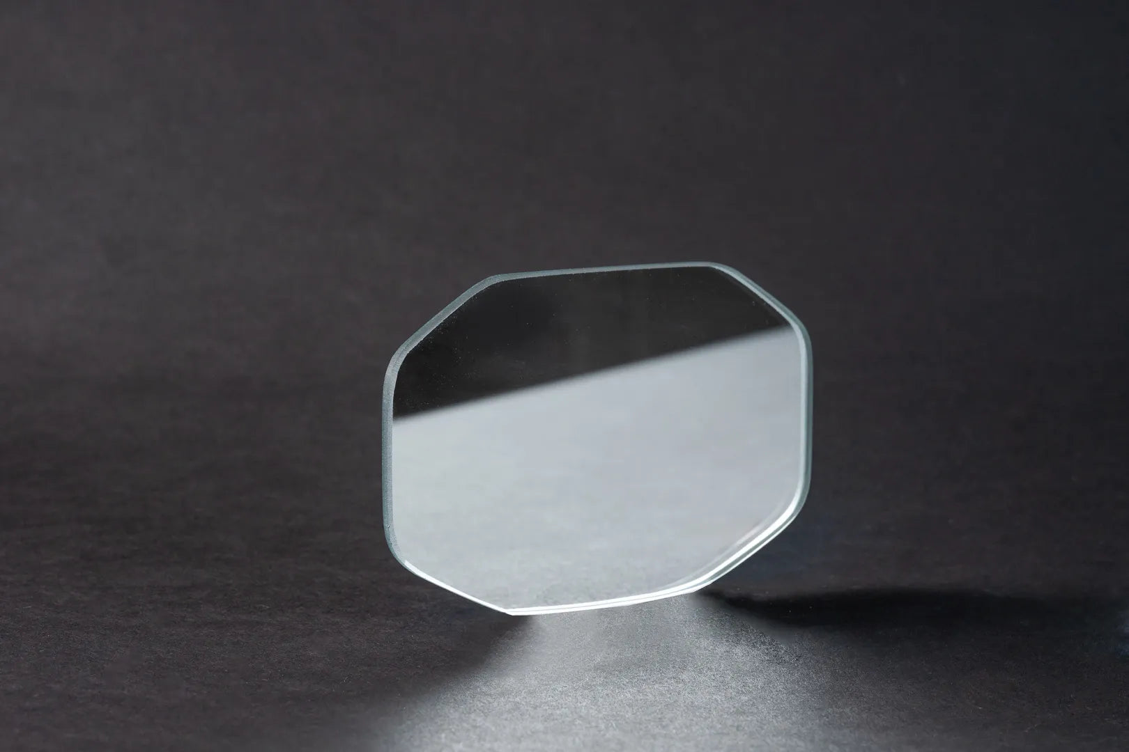 Eine einzelne, transparente, achteckige Glaslinse steht aufrecht auf einer dunklen, strukturierten Oberfläche, unter der sich weiche Schatten und Reflexionen befinden, die an die Klarheit von Oberflächenspiegeln (Weißglas-Basis) erinnern.